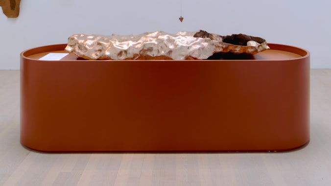 A metal sculpture sits centred on top of a cylindrical oval bronze-coloured plinth.